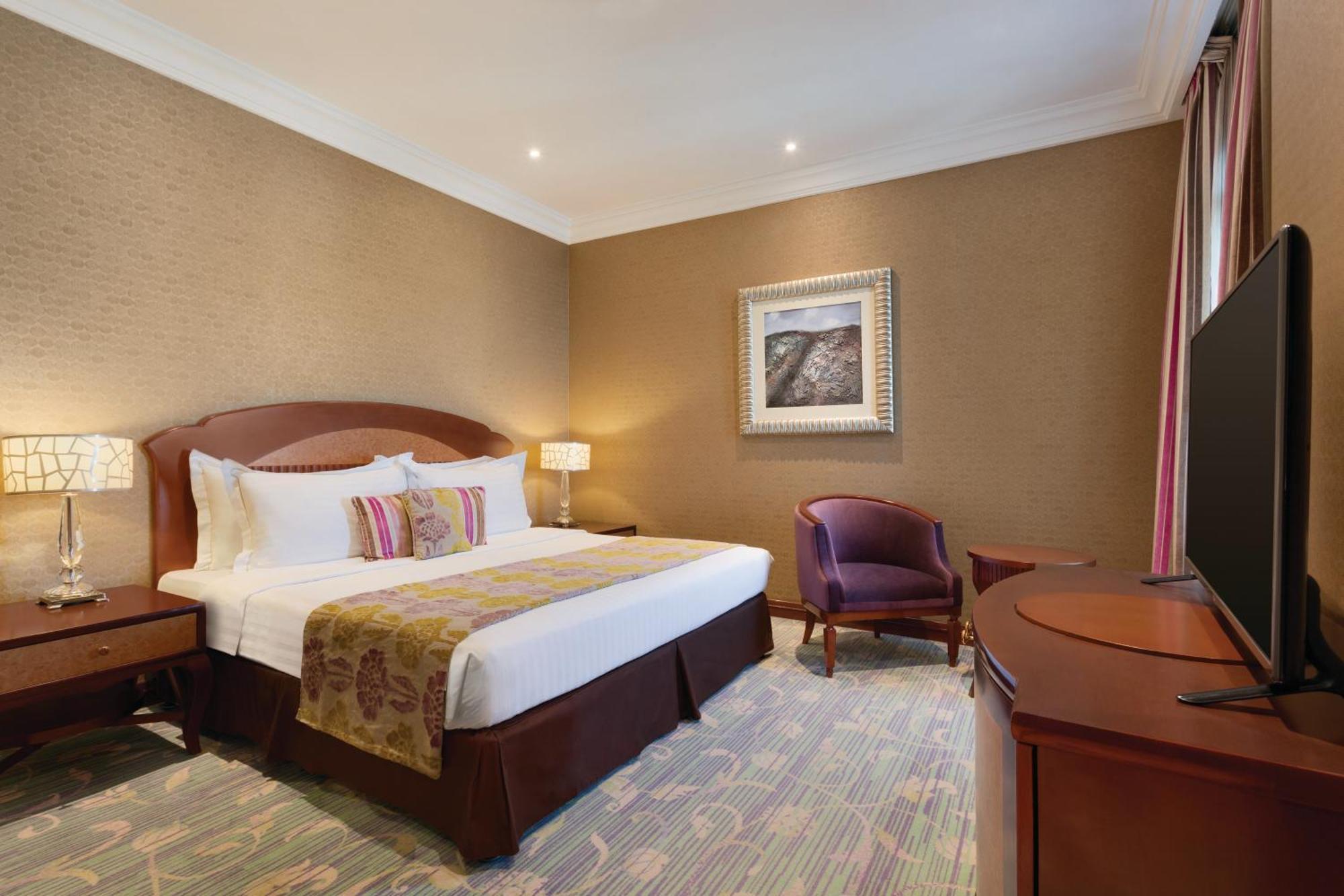 Grand Regency Doha, Trademark Collection By Wyndham Zewnętrze zdjęcie The image shows a hotel room that features a cozy and elegant design. There's a large bed with white sheets and colorful decorative pillows. Next to the bed, there are nightstands with lamps. A comfortable armchair is positioned in the corner, and a 