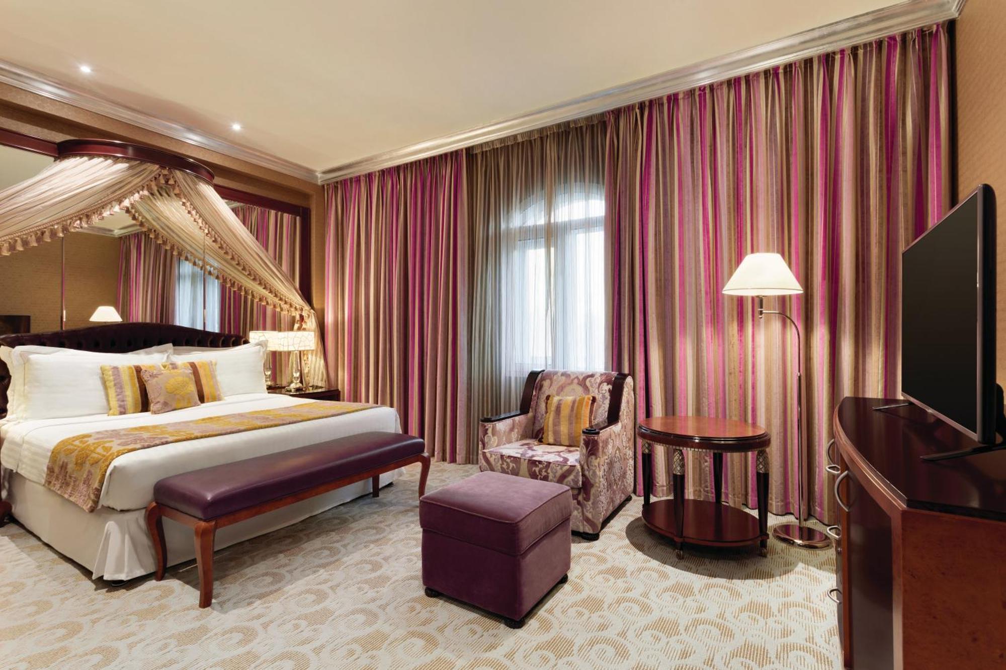 Grand Regency Doha, Trademark Collection By Wyndham Zewnętrze zdjęcie The photo shows a stylish hotel room. It features a large bed with decorative pillows and a draped canopy above it. The room has elegant striped curtains in shades of pink and purple. There is a small seating area with a plush chair and ottoman, alon