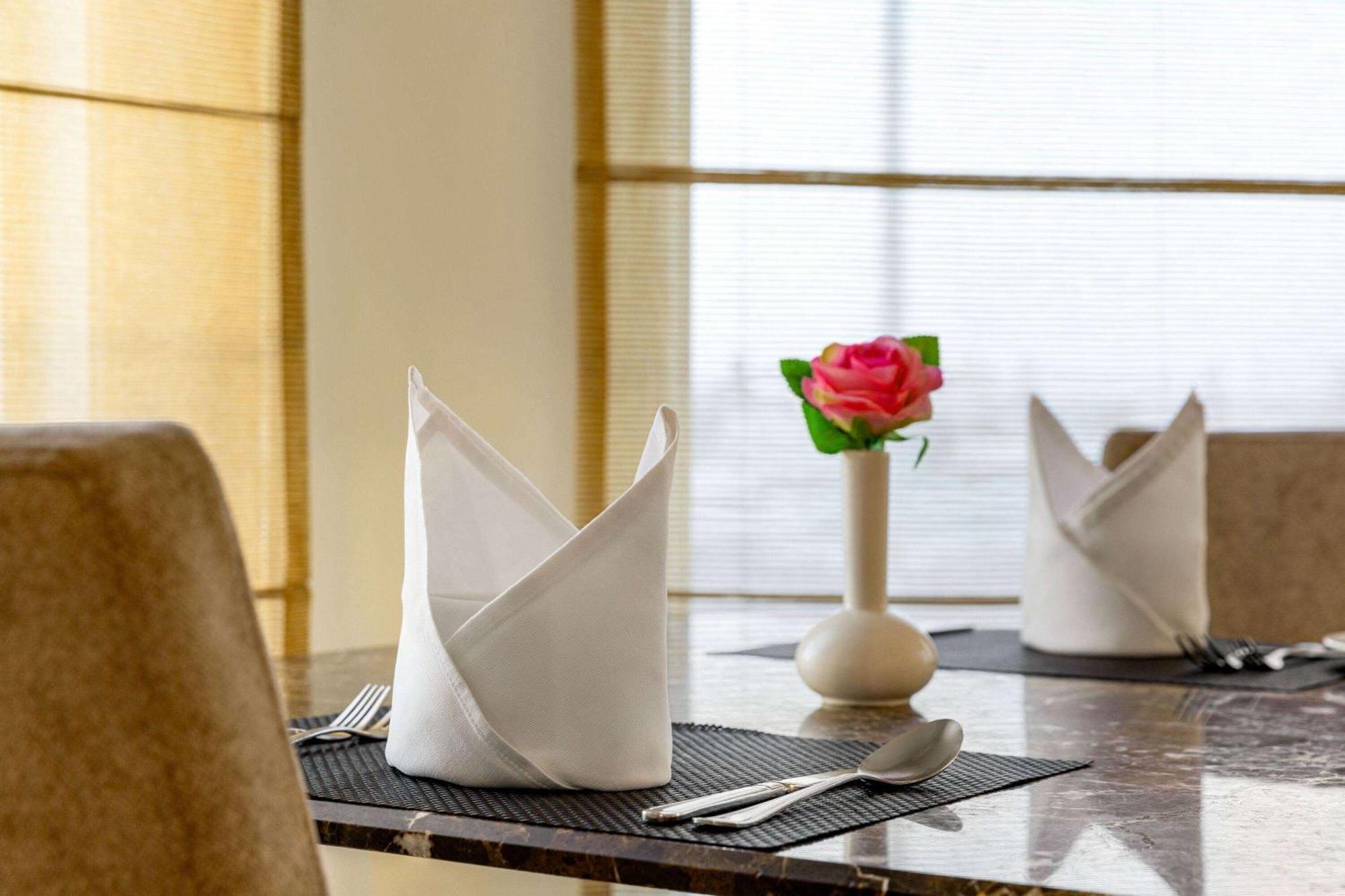 Grand Regency Doha, Trademark Collection By Wyndham Zewnętrze zdjęcie The photo shows a neatly arranged dining table. On the table, there are two cloth napkins folded in a decorative way, one at each place setting. Silverware, including a knife and fork, is placed next to each napkin. In the center of the table, there 