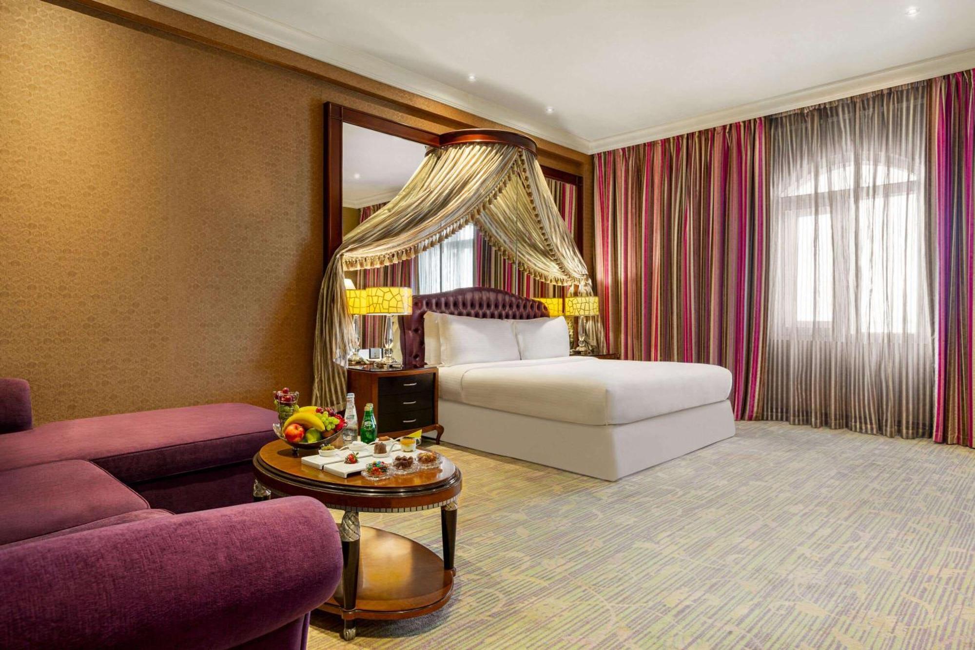 Grand Regency Doha, Trademark Collection By Wyndham Zewnętrze zdjęcie The photo shows a luxurious hotel room featuring a king-sized bed with a decorative canopy. The room is elegantly designed with a mix of rich colors and textures. To the left, there's a plush purple seating area with a small round table that displays